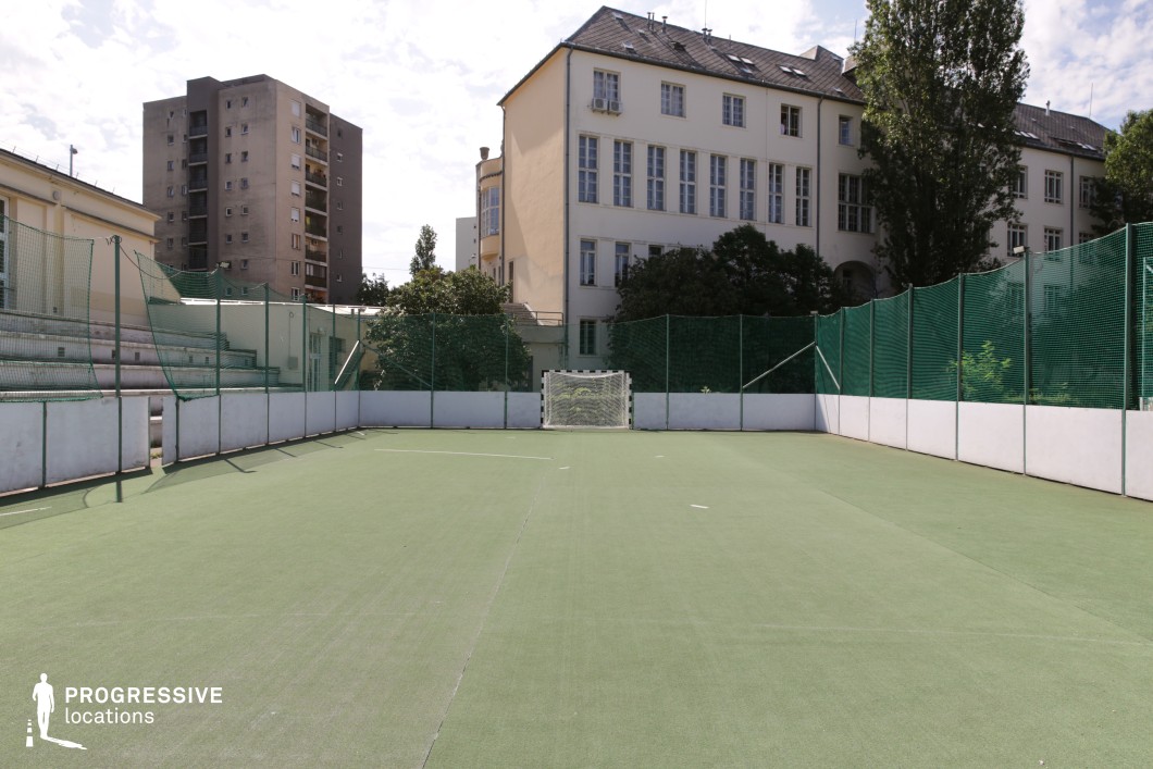 This outdoor sports court features a green artificial turf surface, enclosed by high protective netting and white barriers, creating an intimate yet functional athletic space. A small goalpost sits at one end, while concrete bleachers provide a modest spectator area. Surrounded by classic institutional architecture and modern residential buildings, the court offers a blend of old and new urban elements. Ideal for coming-of-age dramas, sports films, or gritty social narratives, this location provides a versatile backdrop for themes of competition, teamwork, and personal growth.