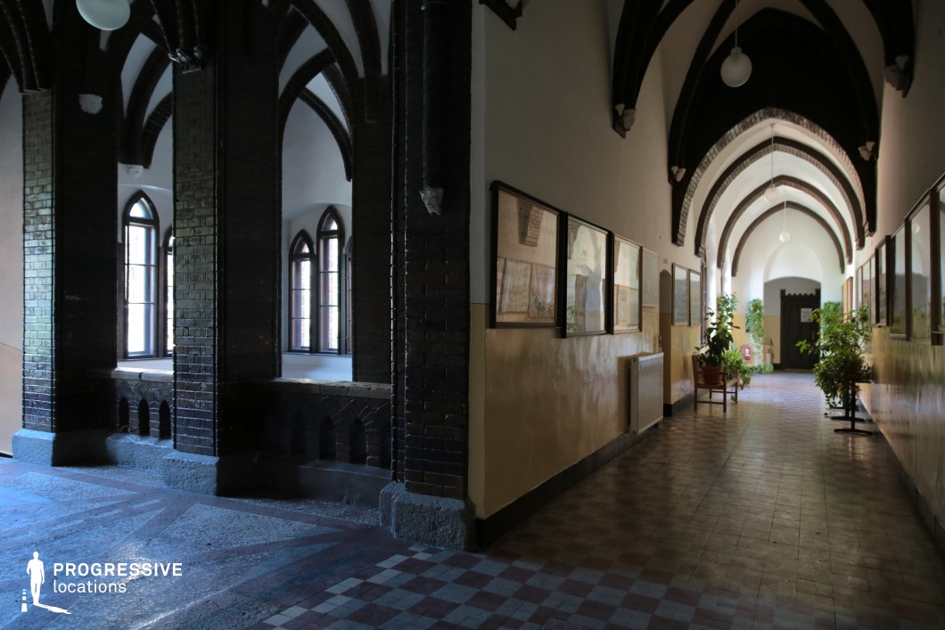 A striking historical corridor with Gothic-style pointed arches and a combination of dark brick and light plaster walls. The space is divided into two sections: one side features large arched windows framed by deep recesses, allowing soft natural light to filter through, while the other side showcases a long, arched hallway adorned with framed artwork and decorative elements. The patterned tile floor and intricate ceiling details enhance the period atmosphere, making this an ideal setting for historical dramas, academic institutions, or atmospheric, mysterious scenes.