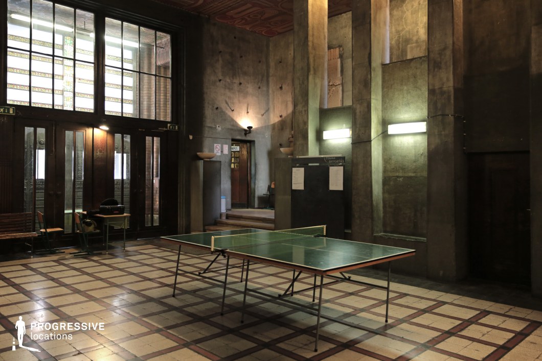 A dimly lit, industrial-style interior a checkered tile floor. The tall concrete walls, adorned with worn textures and aged patina, create a raw, utilitarian aesthetic. Large glass-paneled doors with intricate patterns allow some natural light to enter, contrasting with the artificial glow of wall-mounted tube lights. The space includes retro wooden furniture, a chalkboard with notices, and raised platforms leading to doorways, adding depth and structure. This atmospheric location is ideal for settings requiring a gritty, underground, or vintage sports hall ambiance.