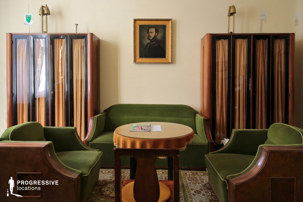 This intimate lounge exudes a mid-century aristocratic charm with its deep green velvet seating, wooden-framed armchairs, and a round table draped in a decorative cloth. Two tall cabinets with pleated curtains flank the space, adding symmetry and an air of formality. The subdued beige walls are adorned with a classical portrait in a gilded frame, reinforcing the vintage aesthetic. The combination of warm wood tones, patterned carpeting, and soft lighting creates a refined yet slightly aged atmosphere. Ideal for period dramas, political thrillers, or introspective character-driven films, this location offers a distinguished setting for dialogue-heavy or historically inspired scenes.