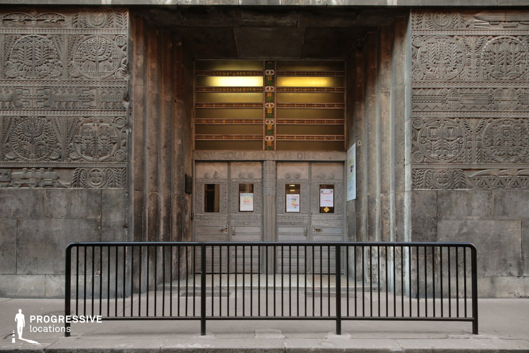 This imposing entrance is framed by massive stone walls adorned with intricate geometric and symbolic relief carvings, creating a bold and historic architectural statement. The heavy metal doors, embellished with elaborate patterns, are topped by a Latin inscription, enhancing the institutional grandeur. Above the doors, a row of stained glass elements adds a subtle decorative contrast to the otherwise austere stone structure. The overall design evokes strength, authority, and permanence, making this location ideal for historical dramas, dystopian thrillers, or academic and governmental settings that require a sense of prestige and mystery.