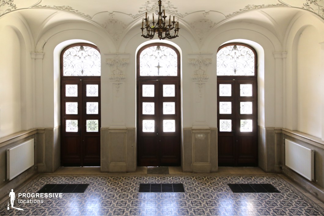 This stately entrance hall features high arched doorways with intricate wrought-iron and frosted glass panels, framed by sculpted stucco detailing. The patterned tile flooring enhances the classical aesthetic, while the chandelier and soft natural light add warmth and sophistication. The symmetrical composition, with rich wooden doors and decorative moldings, evokes a timeless European elegance. Ideal for historical dramas, institutional settings, or mystery films, this location provides an atmospheric backdrop suited for themes of prestige, secrecy, or transition between worlds.