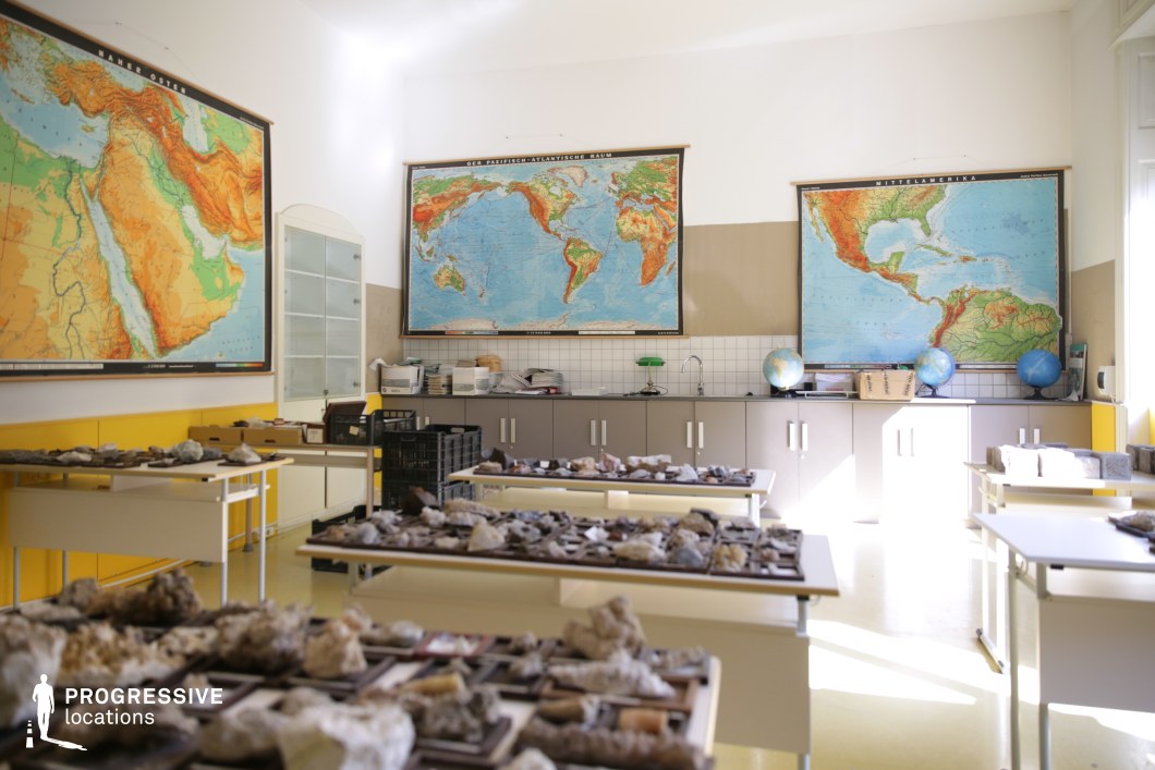 This well-lit geography classroom features large, vintage-style wall maps depicting different regions of the world, creating an academic and exploratory atmosphere. The space is furnished with simple tables displaying an extensive collection of geological specimens, enhancing its educational setting. A row of cabinets along the back wall holds additional materials, while globes and reference materials add to the immersive learning environment. The combination of warm yellow tones and natural light gives the room a nostalgic yet functional feel. Ideal for documentary-style shoots, academic settings, or coming-of-age films, this location provides an authentic backdrop for themes of discovery, education, and curiosity.