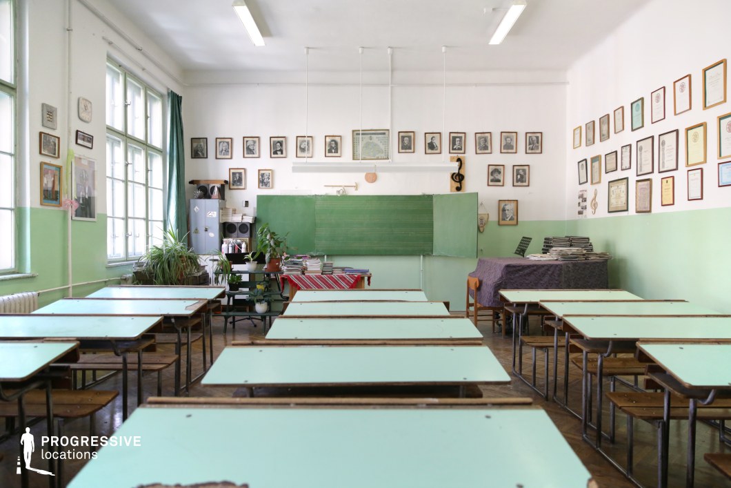 This historic classroom exudes a nostalgic, mid-century academic atmosphere with its pastel green and white walls, worn wooden desks, and classic green chalkboard featuring musical notation. The walls are adorned with framed portraits, certificates, and educational memorabilia, reinforcing the scholarly setting. Large windows allow natural light to flood the space, softened by heavy green curtains. Potted plants and neatly stacked books add a sense of traditional academia. The room’s aged details, including scuffed desks and parquet flooring, make it an ideal setting for period dramas, coming-of-age stories, or films exploring themes of education, history, or nostalgia.