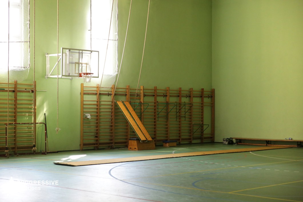 This traditional indoor gymnasium features a pastel green color scheme with high walls, wooden climbing frames, and a suspended basketball hoop. The floor is marked with various sports lines, while gymnastic ropes and angled wooden boards add to the vintage athletic aesthetic. Sunlight streams through tall windows, casting a warm glow on the space and emphasizing the worn textures of the sports equipment. The simple, utilitarian design makes this location ideal for filming nostalgic school settings, physical training sequences, or scenes in sports dramas and coming-of-age films.