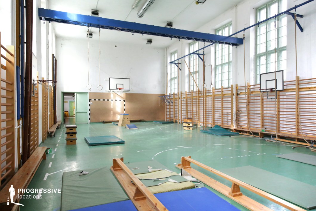 This spacious gymnasium features high ceilings, large industrial-style windows, and a worn green sports floor, creating a nostalgic yet functional atmosphere. Wooden wall bars, hanging gymnastic rings, and climbing ropes add to the traditional school gym aesthetic, while faded mats and training equipment enhance its authentic, slightly aged appearance. The pale walls and soft natural light filtering through the tall windows provide a neutral, airy backdrop, making the space visually dynamic. Ideal for sports-themed scenes, period dramas, or coming-of-age films, this location lends itself well to narratives centered on competition, perseverance, or nostalgia.