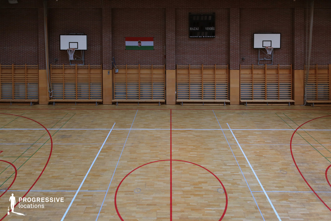 This traditional indoor gymnasium features a polished wooden floor marked with red, white, and green sports lines, set against a backdrop of exposed brick walls. Two basketball hoops are mounted on opposite ends above wooden climbing frames, reinforcing the athletic functionality of the space. A digital scoreboard is positioned centrally, adding to the competitive ambiance. The warm tones of the wood contrast with the industrial brick, creating a balanced and timeless aesthetic. This location is ideal for filming sports dramas, school competitions, or training montages in coming-of-age, motivational, or documentary-style productions.