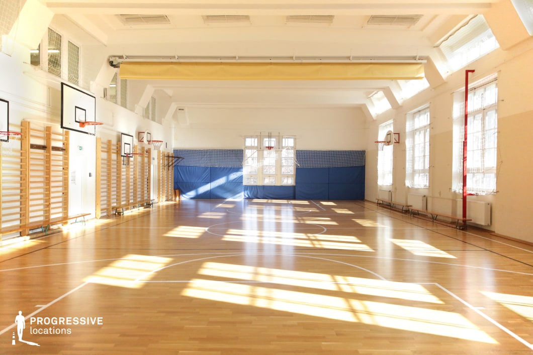 This spacious gymnasium is bathed in natural light, with large windows casting geometric shadows across the polished wooden floor. The high white ceiling features exposed beams and ventilation, contributing to the open and airy feel. Basketball hoops are mounted on the walls, complemented by wooden climbing bars and padded blue wall sections for safety. The clean design, warm tones, and bright atmosphere make this an ideal setting for school sports scenes, training montages, or motivational film sequences in drama, sports, or coming-of-age genres.