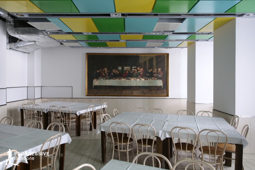 This distinctive dining hall features a large framed reproduction of The Last Supper, prominently displayed against a neutral-toned wall, creating a striking focal point. The space is furnished with simple wooden chairs and square tables draped in white tablecloths, reinforcing a traditional cafeteria setting. The ceiling incorporates colorful geometric panels and exposed ventilation, adding an unexpected modern-industrial contrast to the otherwise subdued interior. This location is ideal for artistic, surreal, or philosophical film projects, as well as dramas or dark comedies, offering a visually intriguing mix of classic and contemporary design elements.