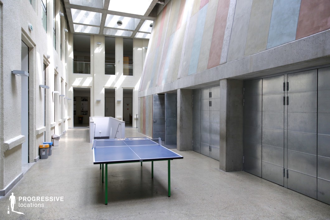 A spacious, modern atrium featuring a high ceiling with a skylight that allows natural light to flood the area. The architecture blends raw concrete surfaces with sleek metallic doors and colorful, pastel-toned wall panels, adding a subtle contrast to the industrial aesthetic. A blue table tennis setup in the foreground introduces a recreational element to the structured environment. The open-plan space is lined with textured white walls and large rectangular windows, providing depth and a contemporary feel. Ideal for filming academic, corporate, or institutional settings with a minimalist and industrial touch.