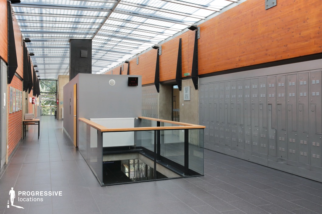 This modern school corridor blends industrial and minimalist design elements, featuring exposed metal lockers, warm wooden paneling, and structural black metal beams. A glass-paneled railing surrounds a stairwell, adding to the open and spacious feel, while a skylight roof allows natural light to illuminate the sleek tiled floor. Concrete walls and metal accents contribute to the raw, utilitarian aesthetic, while a digital clock and signage reinforce the institutional function of the space. This location is ideal for filming contemporary school or academic settings, youth dramas, or sci-fi thrillers requiring a structured yet atmospheric backdrop.