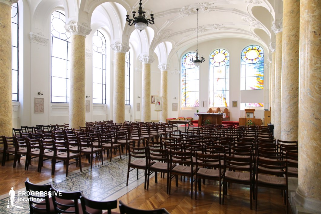 This grand ceremonial hall features towering marble columns, intricate stucco detailing, and expansive arched windows adorned with vibrant stained glass. Rows of wooden chairs face a raised platform, enhancing the formal and institutional ambiance. The polished parquet flooring and decorative tile inlays add elegance, while the high ceilings and chandeliers create a luminous, airy atmosphere. The blend of neoclassical and ecclesiastical elements makes this location ideal for historical dramas, academic or religious ceremonies, or films requiring a prestigious and reverent setting.