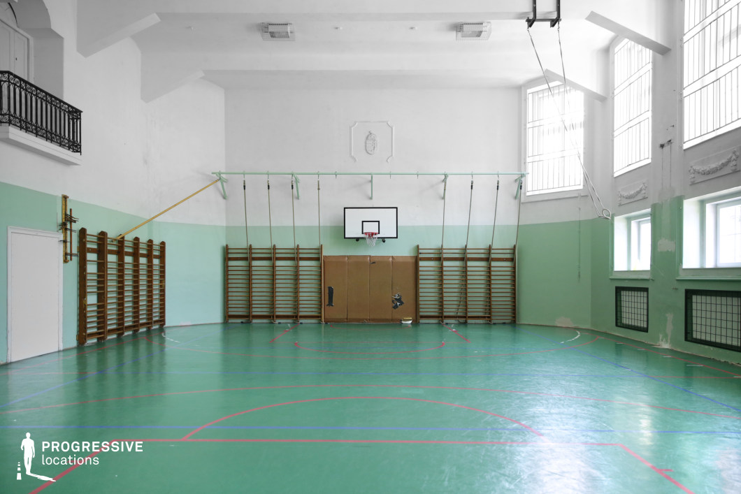 This gymnasium features a retro aesthetic with pastel green and white walls, high arched ceilings, and large windows with decorative molding that allow for abundant natural light. The polished green sports floor is marked with red and blue lines, complementing the wooden climbing frames and gymnastic ropes hanging from the walls. A basketball hoop is centrally positioned above padded wall panels, adding to the institutional character of the space. A small balcony with an ornate wrought-iron railing enhances the classical architectural elements. The combination of aged surfaces and vintage sports equipment makes this location ideal for filming nostalgic, school-themed, or dramatic scenes, particularly in coming-of-age, historical, or psychological thriller genres.
