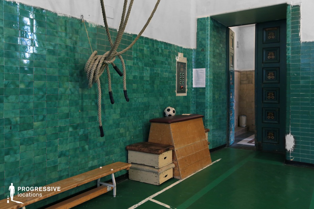 This vintage gymnasium corner features deep green glazed tiles covering the walls, creating a bold and nostalgic atmosphere. Thick gymnastic ropes hang from the ceiling, while aged wooden exercise equipment, including a vaulting box and bench, add to the classic sports hall aesthetic. The doorway, framed by the same green tiles, leads to another section of the building, reinforcing the institutional character. The worn surfaces and distinct mid-century design make this an excellent filming location for period dramas, sports history documentaries, or psychological thrillers requiring a retro, slightly eerie atmosphere.
