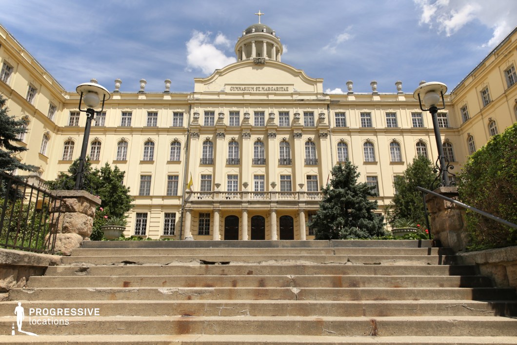 A stately neoclassical building with an ornate façade, featuring tall arched windows, decorative columns, and a central dome crowned with a cross. The grand staircase leading up to the entrance exhibits signs of age, adding to the historical charm of the site. Flanked by lush greenery and vintage-style street lamps, the structure exudes an air of academic prestige and European elegance. The warm yellow hue of the building contrasts beautifully with the clear blue sky, making it a visually striking location ideal for historical documentaries, period dramas, or luxury branding campaigns.