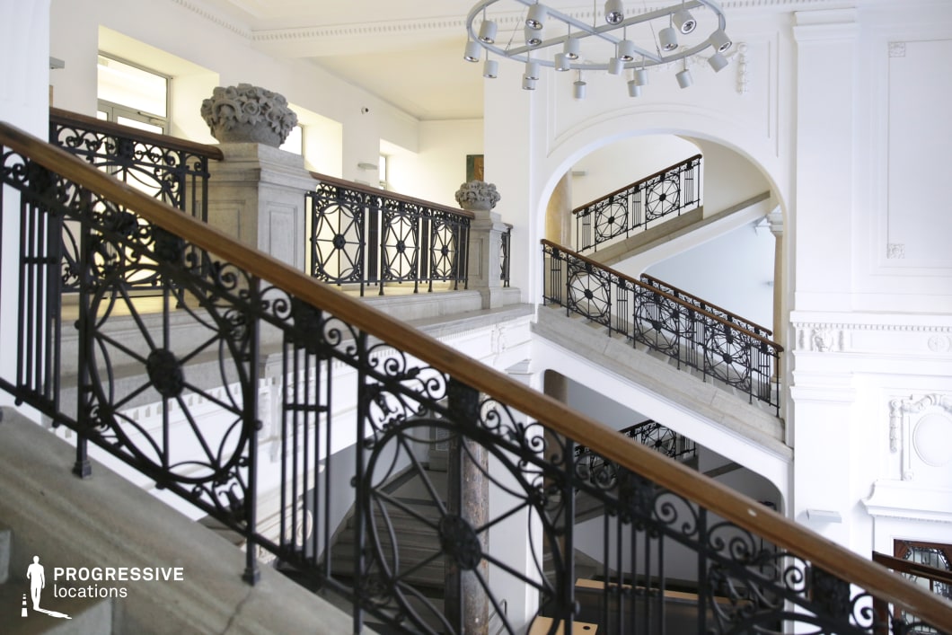 This grand, multi-level staircase embodies classical elegance with its intricate wrought iron railings and finely carved stone balustrades. The polished wooden handrails add warmth to the predominantly white interior, which is accentuated by high ceilings, arched openings, and decorative molding. A contemporary circular light fixture contrasts with the historical charm, blending old-world sophistication with modern design elements. The space is well-lit by natural light streaming through large windows, emphasizing the refined architectural details. Ideal for film productions requiring a stately academic setting, historical ambiance, or upscale institutional aesthetics.