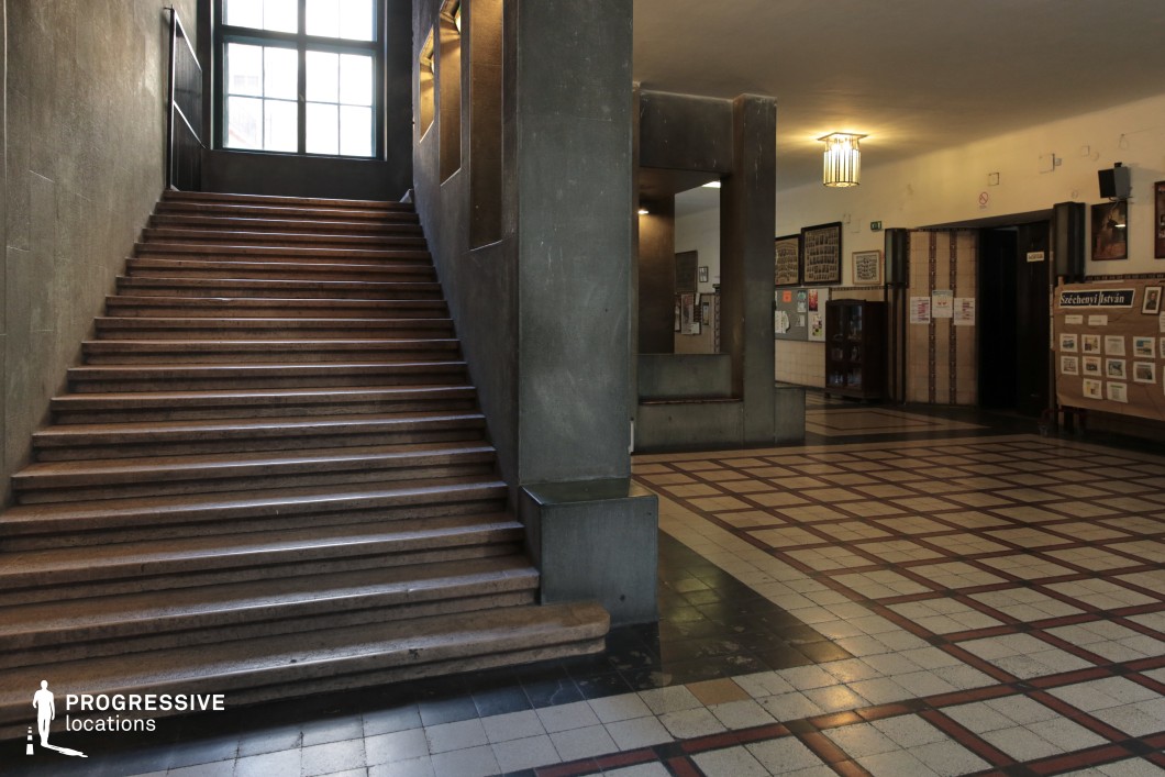 This imposing interior features a wide stone staircase with dark, textured walls, leading to a large window that filters in moody natural light. The checkered tile flooring and geometric architectural details contribute to the austere, institutional ambiance. The surrounding hallway, lined with historical displays and bulletin boards, enhances the sense of academia or authority. The space’s structured symmetry, cool tones, and heavy materials make it an ideal setting for historical dramas, noir thrillers, or institutional films exploring themes of power, control, or mystery.