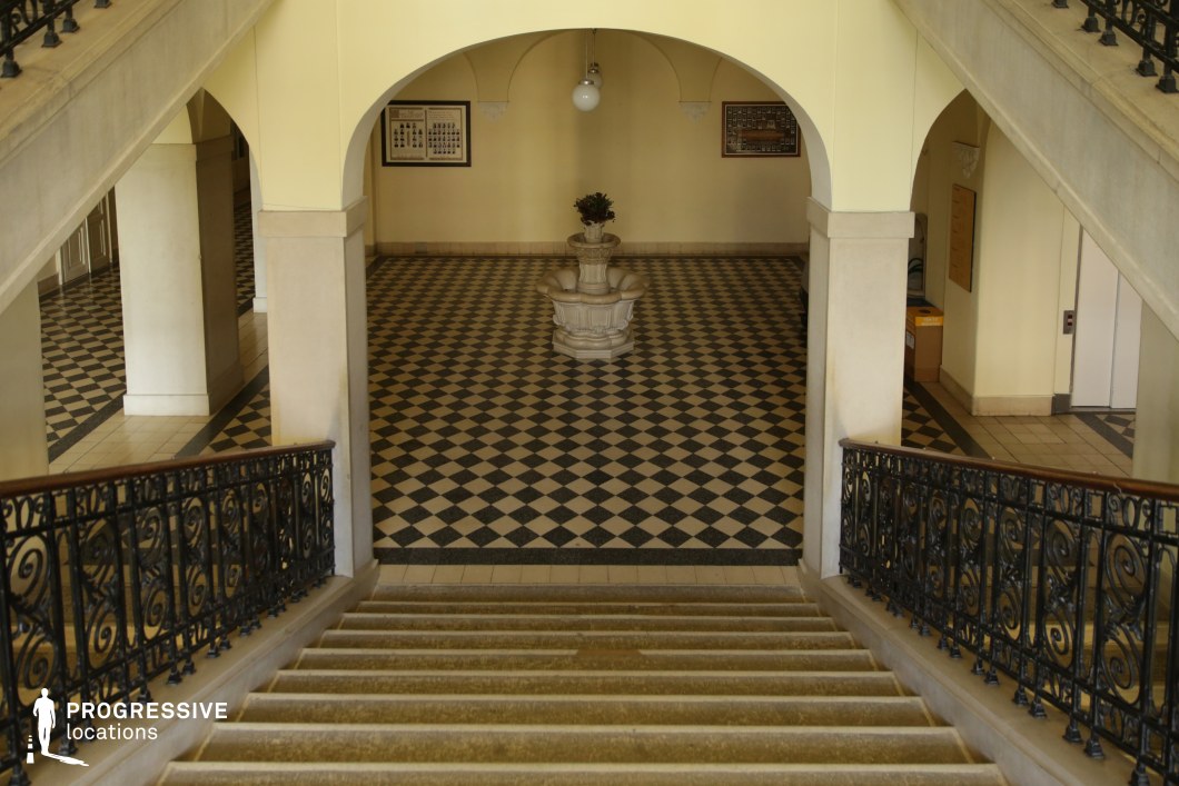 This elegant interior features a sweeping staircase with ornate wrought-iron railings leading to a checkered marble-floored hall. The classical design is emphasized by smooth archways, cream-colored walls, and a central decorative fountain that adds a refined focal point. The symmetry of the space, along with its warm lighting and high ceilings, enhances its grandeur and historical ambiance. Ideal for period dramas, institutional settings, or mystery narratives, this location conveys sophistication, authority, and timeless elegance, making it suitable for scenes set in academies, government buildings, or aristocratic estates.