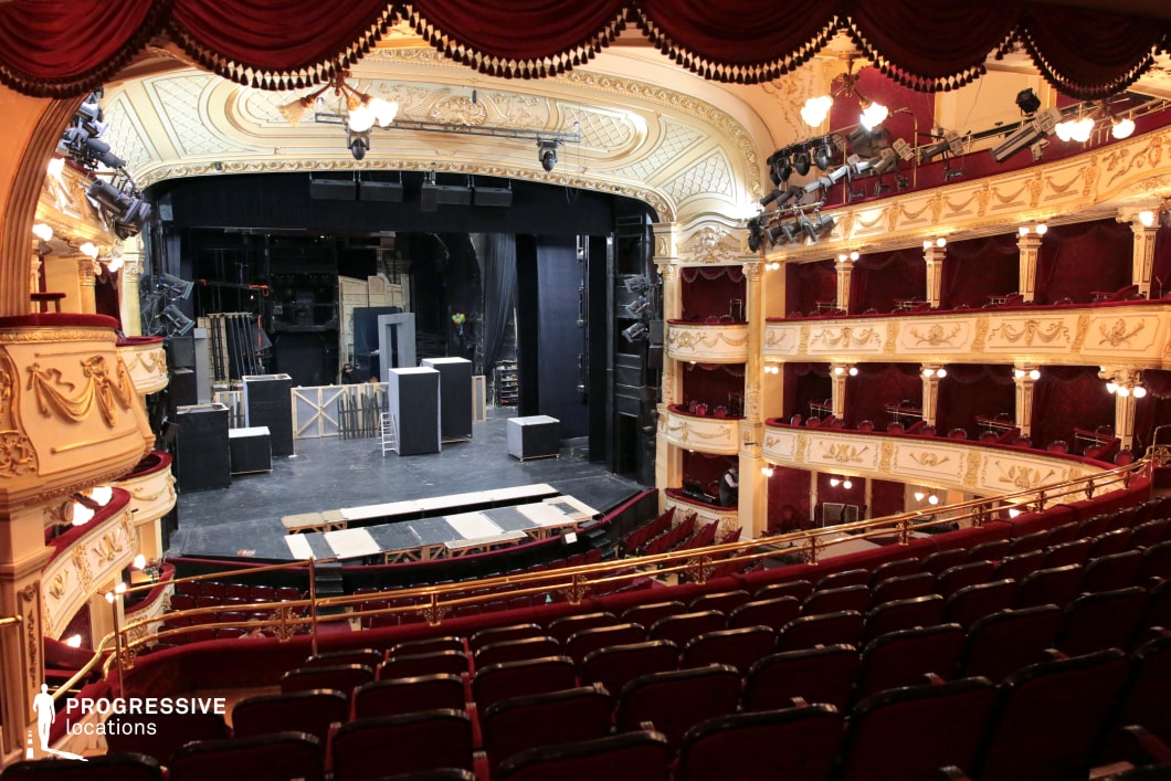 This grand theater auditorium features an opulent design with intricate gold detailing, rich red velvet seating, and elegantly curved balconies. The high, vaulted ceiling is adorned with elaborate moldings, enhancing the classical European aesthetic. The plush seating and decorative box seats evoke a sense of timeless grandeur, making this venue ideal for period film productions, high-end performances, or luxury event settings. The architectural splendor and theatrical atmosphere create a visually stunning and immersive cinematic backdrop.