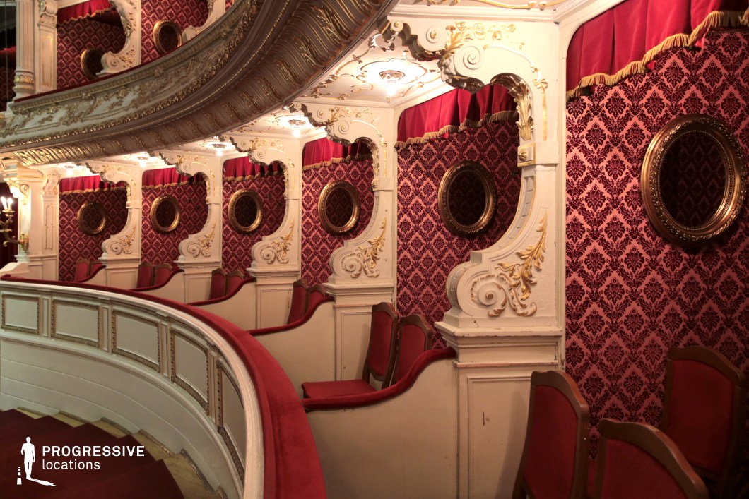 This luxurious theater balcony features intricate baroque-style detailing with gilded embellishments, white carved moldings, and rich red velvet drapery. The damask-patterned wall coverings and round gold-framed mirrors enhance the opulent, old-world aesthetic. The plush red velvet seats, arranged in intimate boxes, create an exclusive and aristocratic atmosphere. The curved railing and softly lit ceiling details add to the grandeur of the space. This setting is ideal for period dramas, historical romances, or suspenseful thrillers requiring a setting of elegance, wealth, and theatrical intrigue.