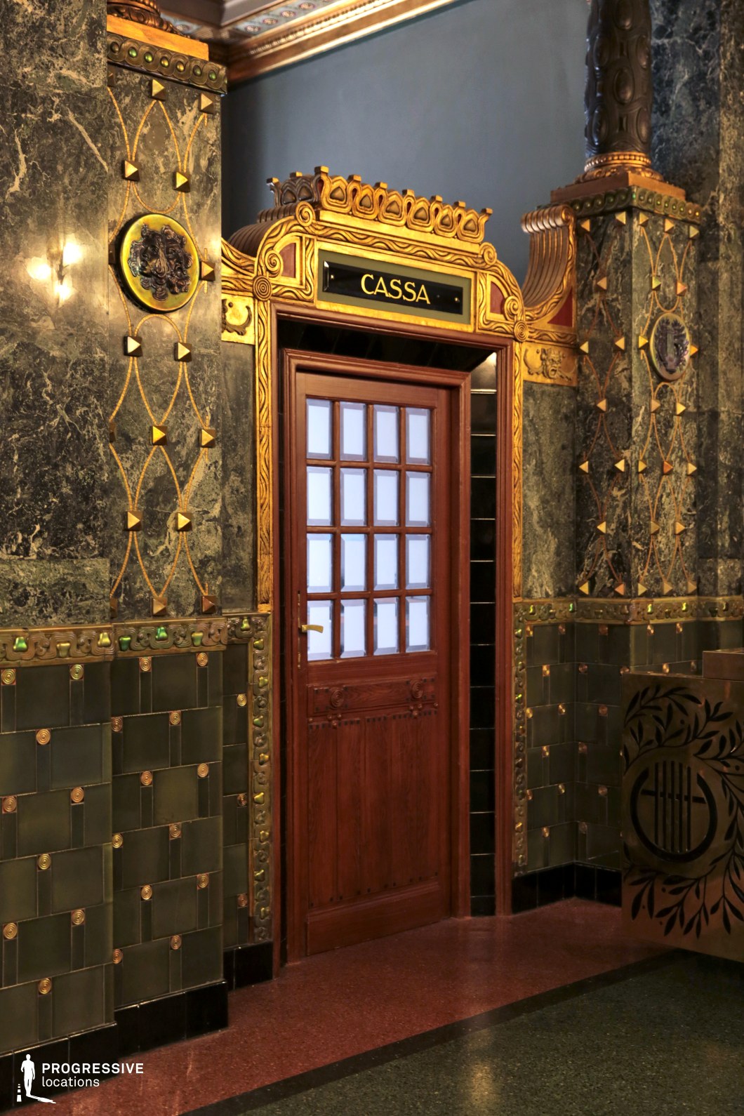 A beautifully detailed ticket booth entrance framed by rich Art Nouveau design elements. The wooden door, with its frosted glass panes, is set within a gilded archway adorned with intricate geometric patterns and organic motifs. The surrounding walls feature dark green marble with gold embellishments and ceramic tilework, adding texture and depth. Small sculptural details and decorative elements enhance the historical character of the space. This setting is ideal for period films, classic European settings, or sophisticated scenes requiring an elegant and nostalgic ambiance.