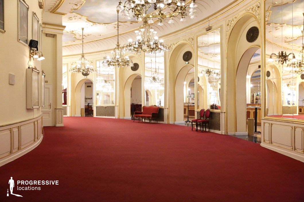 A luxurious theater corridor which doubles as a ballroom with a grand red carpet, intricate gold detailing, and tall mirrored arches that enhance the sense of space and opulence. The ceiling features ornate frescoes framed by curved molding, while multiple crystal chandeliers cast a warm, ambient glow. The symmetrical design, elegant furnishing, and reflective surfaces contribute to a refined, aristocratic atmosphere. This setting is ideal for period dramas, high-society events, or historical romance films, providing a visually stunning backdrop for scenes of grandeur and sophistication.