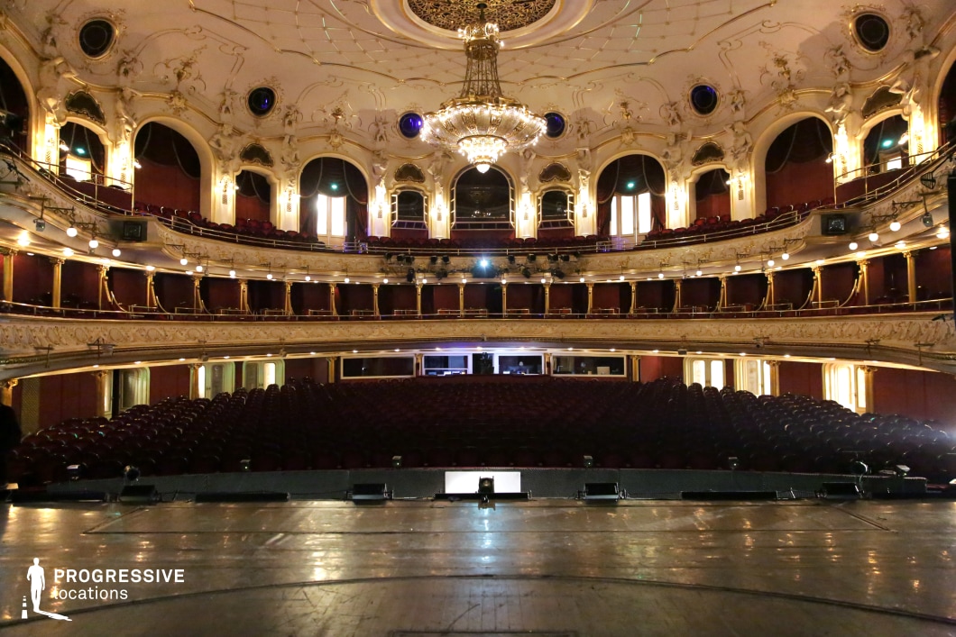 A lavish, historic theater with an opulent multi-tiered balcony arrangement, intricate gold detailing, and deep red velvet accents. The ceiling features elegant sculptural elements and a grand chandelier, casting a warm glow over the ornate architectural design. Arched windows and decorative moldings add a regal charm, enhancing the sense of luxury and prestige. The stage’s perspective emphasizes the immersive depth of the auditorium, making this location ideal for period dramas, operatic performances, and high-society cinematic settings that require a rich, classical atmosphere.