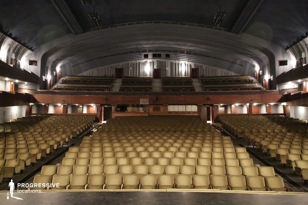 This vast auditorium embodies Soviet-era brutalist architecture with its sweeping curved ceiling, dark muted tones, and rigid geometric structure. Rows of beige seats extend across multiple levels, framed by a stark contrast of wood paneling and industrial concrete. The dim lighting and cavernous space create a subdued yet imposing atmosphere, while the symmetrical layout emphasizes order and functionality. This setting is ideal for dystopian narratives, political thrillers, or historical dramas, offering a strikingly austere backdrop that conveys authority, nostalgia, or institutional grandeur.