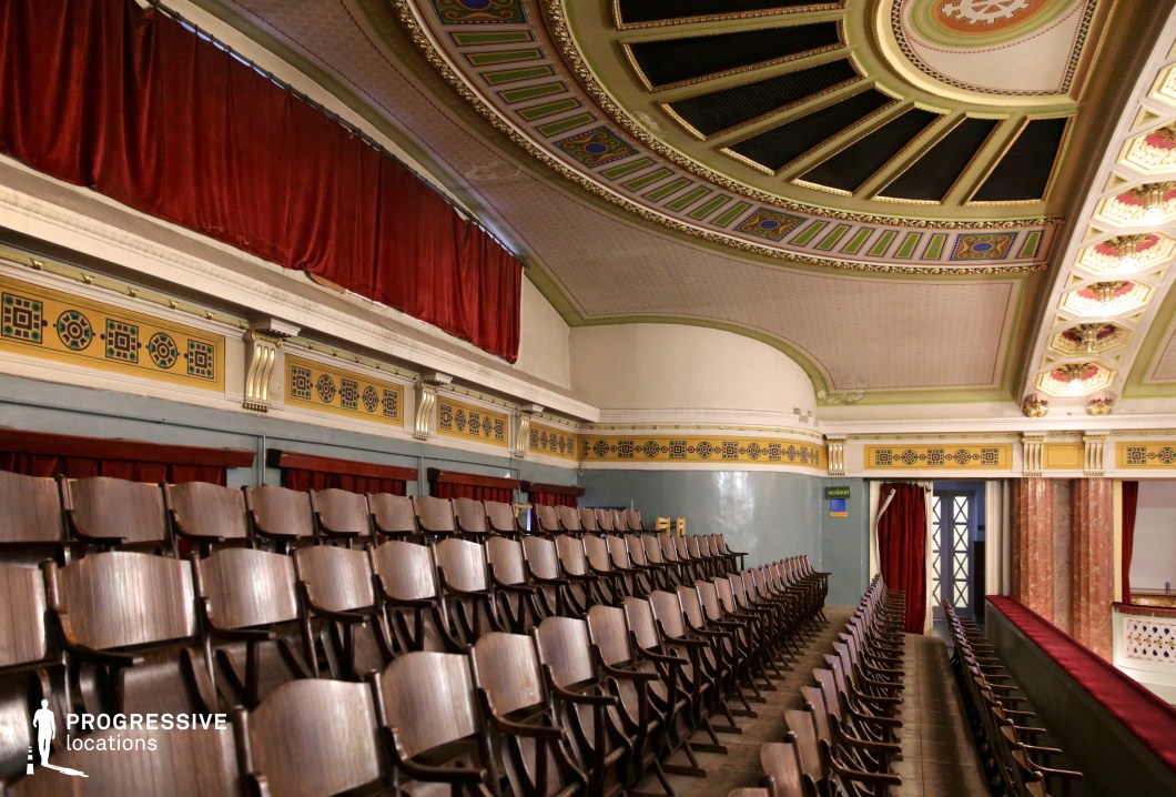This grand theater balcony features rows of vintage wooden seats set against a richly decorated ceiling with intricate geometric and floral patterns. The curved architectural design, highlighted by gold accents and pastel-hued paneling, enhances the historical character of the space. Deep red velvet drapery adds a touch of warmth and sophistication, complementing the classic color palette. The combination of elegant craftsmanship and a slightly aged ambiance makes this setting ideal for period films, political dramas, or historical thrillers requiring a timeless, atmospheric backdrop.