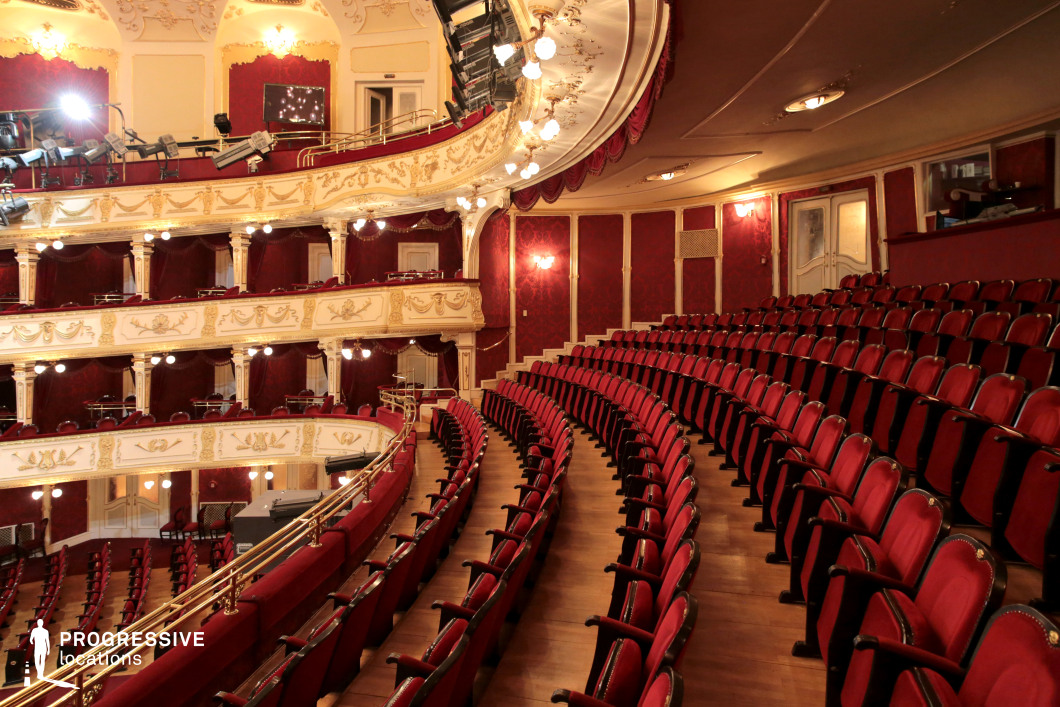 A grand historic theater featuring luxurious red velvet seating, ornate white and gold balconies, and intricately detailed molding. The curved arrangement of seats offers an immersive perspective, while decorative sconces and soft ambient lighting enhance the warm, sophisticated ambiance. The plush red drapery and classical embellishments evoke an old-world elegance, making this venue an ideal setting for period dramas, aristocratic gatherings, or refined theatrical performances requiring a regal and opulent backdrop.