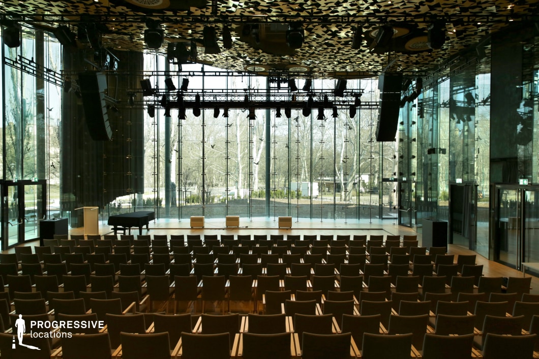 A contemporary concert hall with floor-to-ceiling glass walls, offering an open, airy ambiance and a seamless connection to the surrounding outdoor landscape. The minimalist interior features warm wooden flooring, sleek seating arrangements, and a spacious stage equipped with professional lighting and audio systems. The ceiling is adorned with an intricate geometric pattern, adding a striking visual contrast. The transparent backdrop allows for dynamic natural lighting, making this an ideal location for live performances, high-end conferences, or stylish cinematic scenes that require a blend of modernity and elegance.