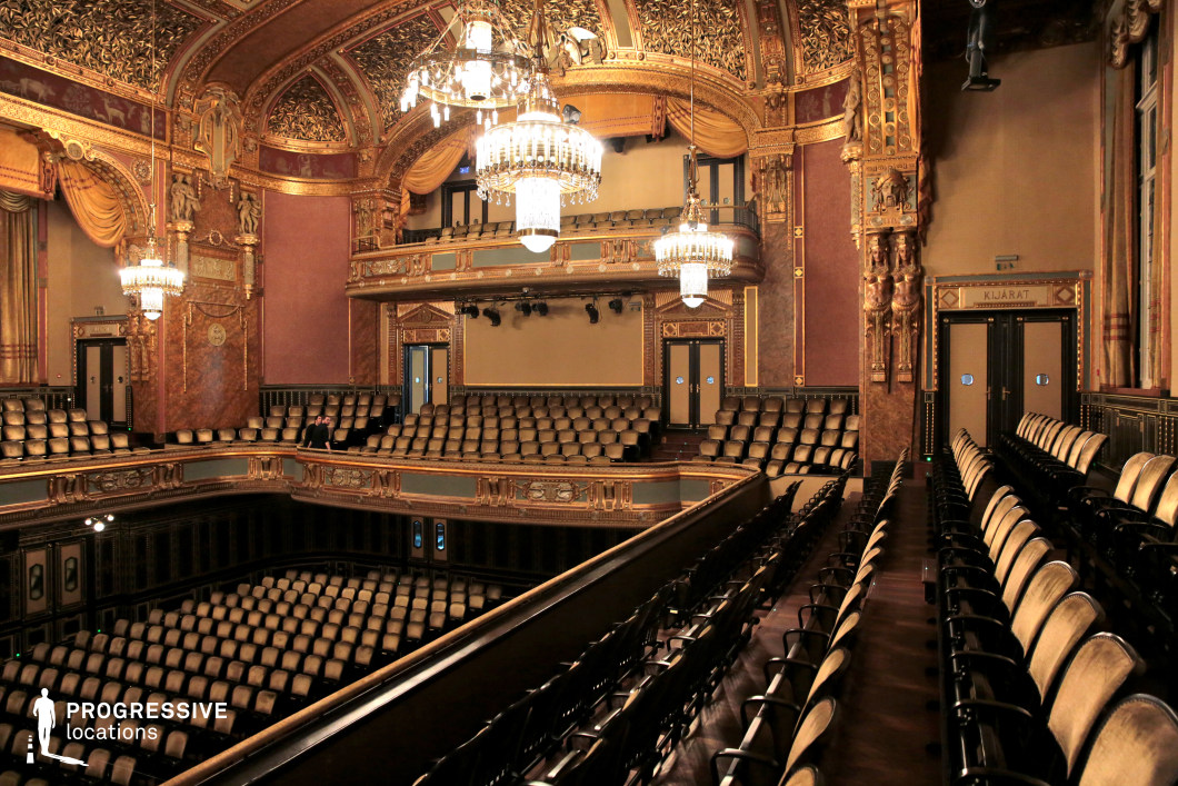 A lavishly decorated theater with a grand multi-tiered seating arrangement, featuring intricate gold embellishments, rich wood paneling, and deep red drapery. The arched ceiling is adorned with elaborate gilded patterns, while large crystal chandeliers provide an elegant illumination. The balconies are supported by intricately carved details, evoking a timeless sense of luxury and sophistication. The overall design blends classical and baroque influences, making this location perfect for period dramas, high-society gatherings, or cinematic performances that require an aristocratic and refined setting.