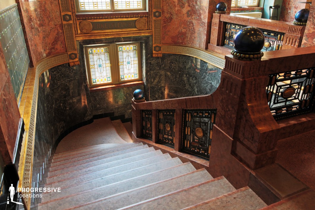 This striking Art Nouveau staircase features richly colored marble, intricate wrought-iron detailing, and stained-glass windows that filter warm, ambient light into the space. The curved handrails and decorative balustrades with geometric patterns contribute to the grandeur, while the polished stone steps enhance the elegance of the design. The combination of gold-accented moldings and deep hues creates a sophisticated, historic ambiance. This setting is ideal for period dramas, mystery thrillers, or classic European-style productions that require an opulent and visually captivating architectural backdrop.