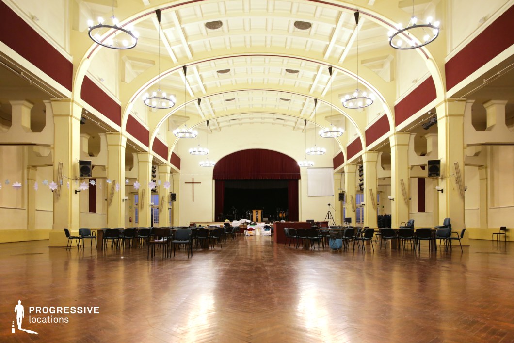 This large assembly hall features high, vaulted ceilings supported by elegant arches, giving the space a grand yet functional aesthetic. The pale yellow walls contrast with deep red accents, including a theater-style stage with a heavy velvet curtain. The polished wooden floor reflects the soft glow of the circular chandeliers, which enhance the sense of openness. The minimal ornamentation and symmetrical structure create a versatile environment. This location is ideal for historical films, political dramas, or institutional settings, providing a timeless and adaptable backdrop for a variety of cinematic narratives.