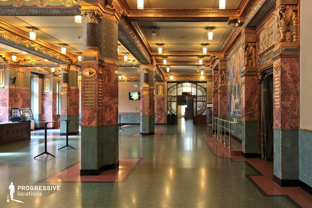 A richly decorated hall featuring intricate architectural details, including marble-clad columns, gilded moldings, and decorative friezes. The Art Nouveau-inspired design incorporates vibrant green and red tones, complemented by geometric patterns and sculptural reliefs. Hanging lantern-style lights provide a warm, ambient glow, while arched doorways with stained glass accents enhance the elegant aesthetic. The polished stone flooring reflects the opulence of the space. This location is ideal for period dramas, historical films, or refined cultural settings requiring a visually striking and sophisticated atmosphere.