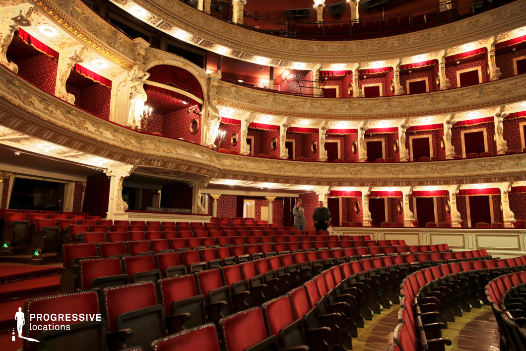 A grand, opulent theater with rich red and gold decor, featuring multiple tiers of private balconies adorned with intricate white sculptural details. The plush red velvet seating and patterned wall coverings create a warm, luxurious ambiance, while ornate lighting fixtures enhance the regal aesthetic. The curved rows of seating provide a dynamic perspective, leading toward the stage. This setting is ideal for period dramas, operatic performances, or high-society film scenes requiring an aristocratic, historically rich backdrop.