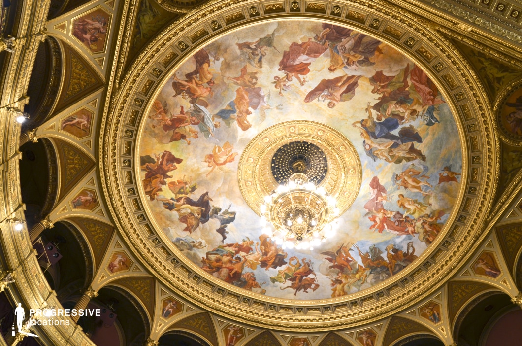 This awe-inspiring ceiling fresco, surrounded by intricate gold detailing, showcases a masterpiece of classical artistry. The scene, filled with dynamic figures in vibrant hues, is illuminated by an opulent central chandelier, enhancing the luxurious atmosphere of the space. Encased within a richly adorned dome, the artwork exudes grandeur and refinement, making it an ideal setting for period films, high-end productions, or luxury-themed editorial shoots. The combination of exquisite craftsmanship and dramatic lighting creates a visually compelling backdrop, perfect for cinematic storytelling and elite event settings.