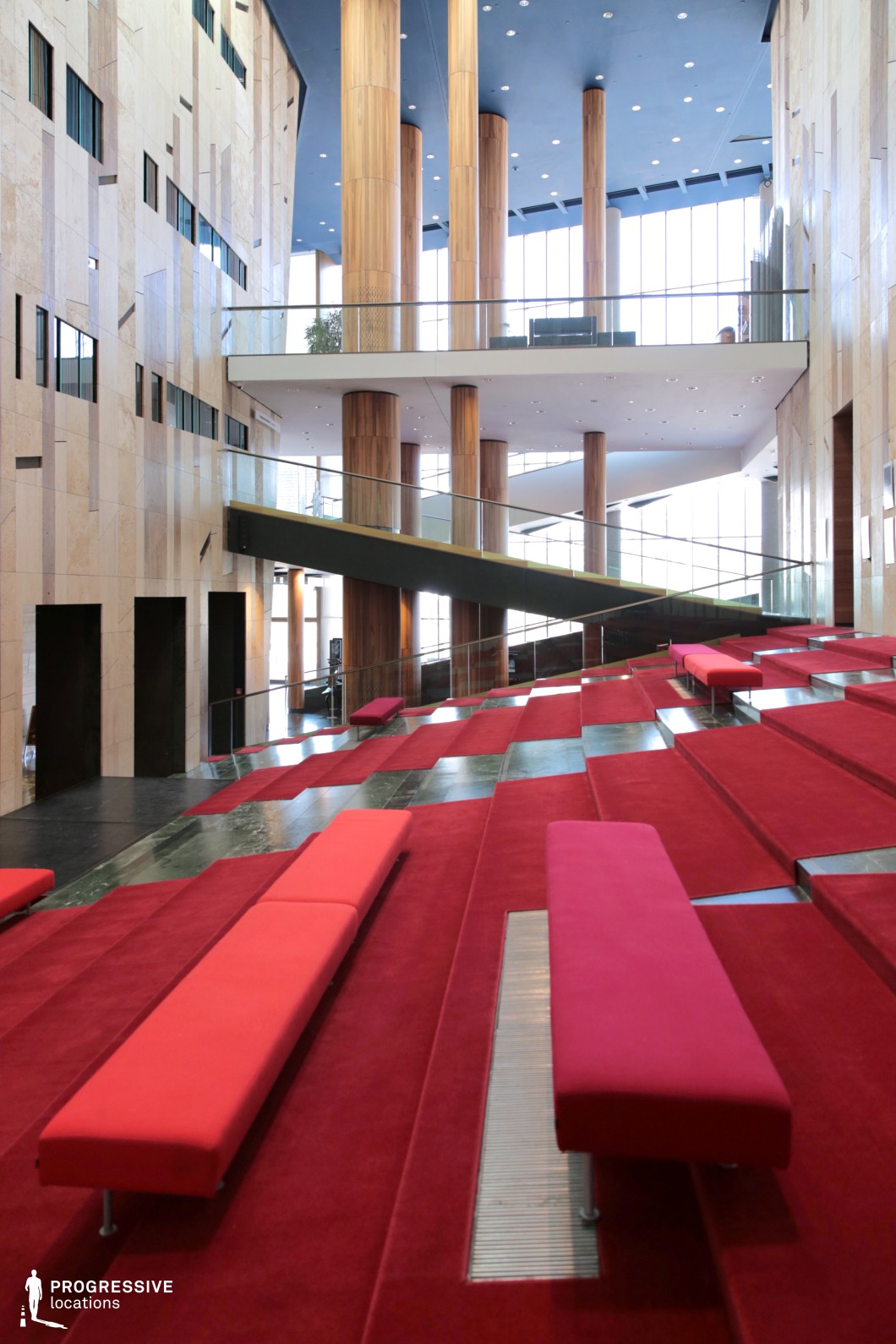 A striking contemporary atrium with an open, multi-level design featuring tiered red-carpeted seating and sleek, minimalistic benches. Tall wooden columns stretch toward the high ceiling, complemented by expansive floor-to-ceiling windows that flood the space with natural light. The floating mezzanine and angular walkways create a dynamic architectural composition, while the mix of stone, wood, and glass adds depth and sophistication. This visually engaging location is ideal for modern corporate settings, futuristic productions, or high-end cultural events requiring a sleek and innovative backdrop.