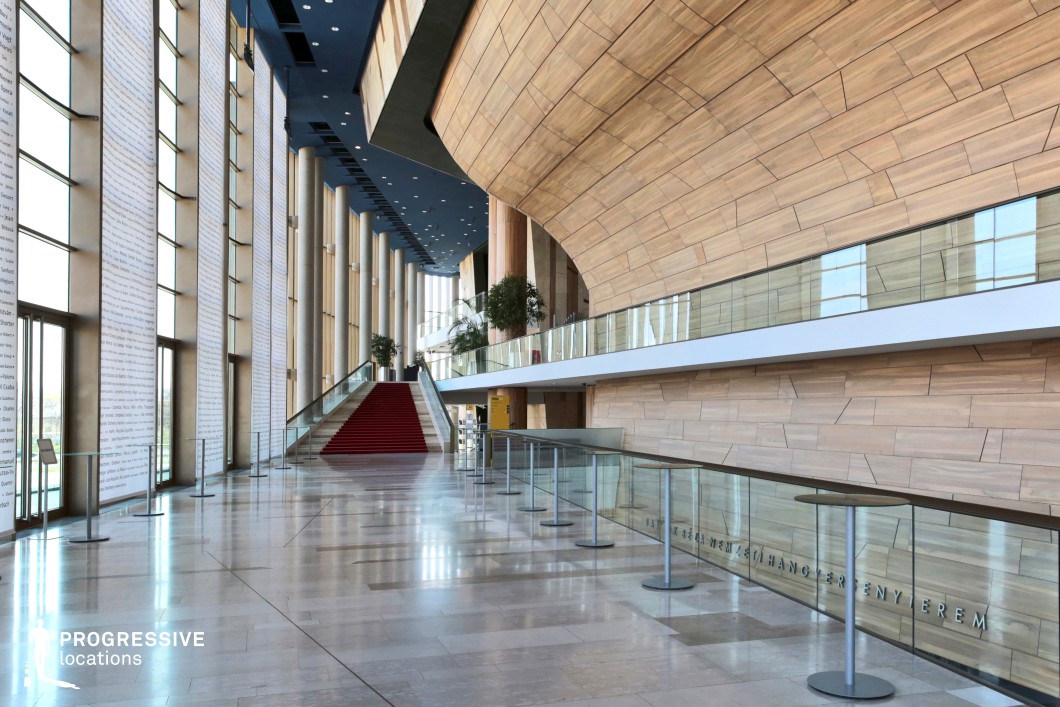 A vast, modern lobby with a sleek, open design, featuring high ceilings, floor-to-ceiling windows, and a dramatic curved wooden wall. The polished stone flooring enhances the spacious feel, while a striking red-carpeted staircase adds a touch of elegance. Vertical illuminated panels line the windows, creating a dynamic visual contrast. The glass railing and mezzanine level offer additional depth and perspective. This sophisticated and airy setting is ideal for high-profile events, corporate scenes, or futuristic and minimalist film productions requiring a stylish, upscale atmosphere.