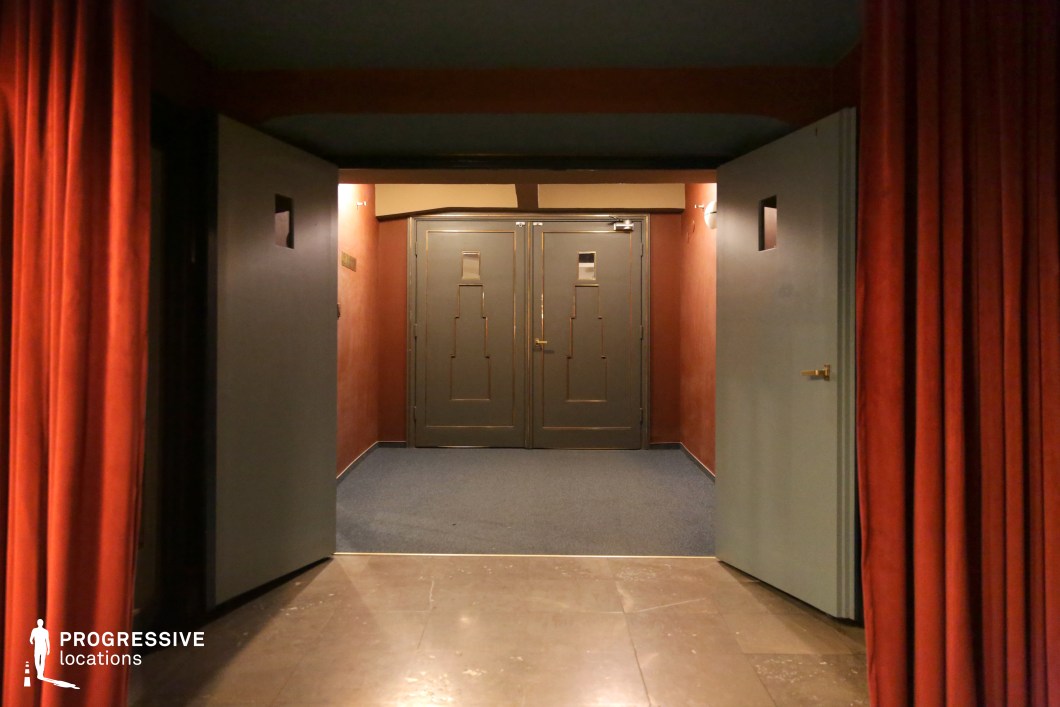 This dimly lit theater or cinema entrance features deep red velvet curtains framing a set of heavy double doors with geometric Art Deco-inspired detailing. The muted color palette, consisting of dark green, rust, and soft lighting, creates an intimate yet slightly mysterious ambiance. The combination of rich textures and subdued lighting enhances the vintage atmosphere. This location is ideal for noir films, suspenseful thrillers, or period dramas requiring a setting with a timeless cinematic allure.