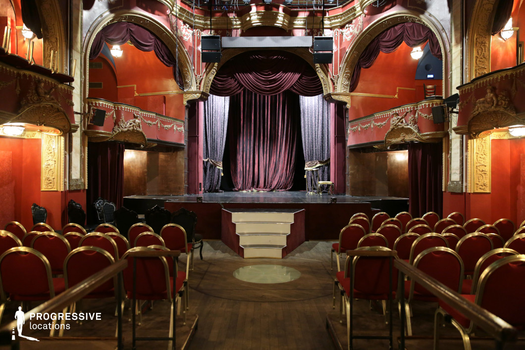 A richly decorated small theater with deep red walls, gold embellishments, and intricate baroque-style moldings. The stage is framed by dramatic velvet curtains, while arched balconies with ornate carvings enhance the historical charm. Rows of red and gold chairs create an elegant yet intimate seating arrangement, while the dark wooden flooring adds warmth. The dim lighting and vintage decor give the space a nostalgic, cabaret-like ambiance. This setting is ideal for period dramas, theatrical performances, or moody, atmospheric film scenes requiring a sense of old-world luxury and intrigue.