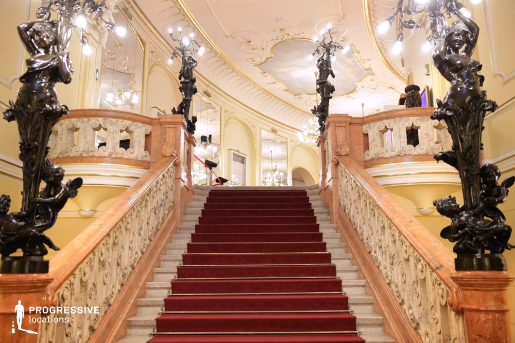 A sweeping, double-sided staircase with a rich red carpet is framed by intricate marble balustrades and adorned with elaborate wrought-iron sculptures featuring cherubs and female figures holding ornate light fixtures. The ceiling showcases elegant gold-accented molding and painted details, reflecting the classical luxury of the space. Large mirrors and chandeliers amplify the grandeur, adding depth and opulence to the scene. This visually striking location is perfect for historical dramas, aristocratic settings, or lavish period films, evoking a sense of prestige, power, and classical elegance.