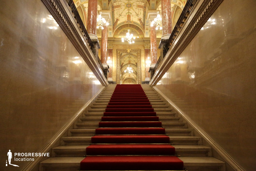 A majestic, wide staircase with a luxurious red carpet ascends toward an opulent hall, framed by polished marble walls with intricate molding. The ceiling features elaborate gold-accented frescoes, complemented by grand chandeliers casting a warm glow. Tall, richly decorated columns with intricate details and a gilded archway add to the grandeur. The symmetrical composition and depth create a cinematic sense of prestige and power. This location is ideal for historical dramas, period films, or scenes depicting aristocratic opulence and formal grandeur.