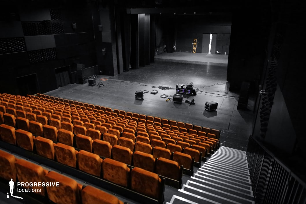 This contemporary black box theater features an expansive stage framed by dark walls and technical rigging, creating a flexible performance space. The tiered seating with plush burnt-orange chairs contrasts with the monochromatic surroundings, adding warmth to the otherwise industrial aesthetic. Scattered technical equipment on the stage suggests a dynamic, adaptable venue suitable for various productions. The minimalist design, exposed structural elements, and functional layout make this an ideal setting for avant-garde performances, experimental films, or high-stakes dramas that require a raw, immersive atmosphere.