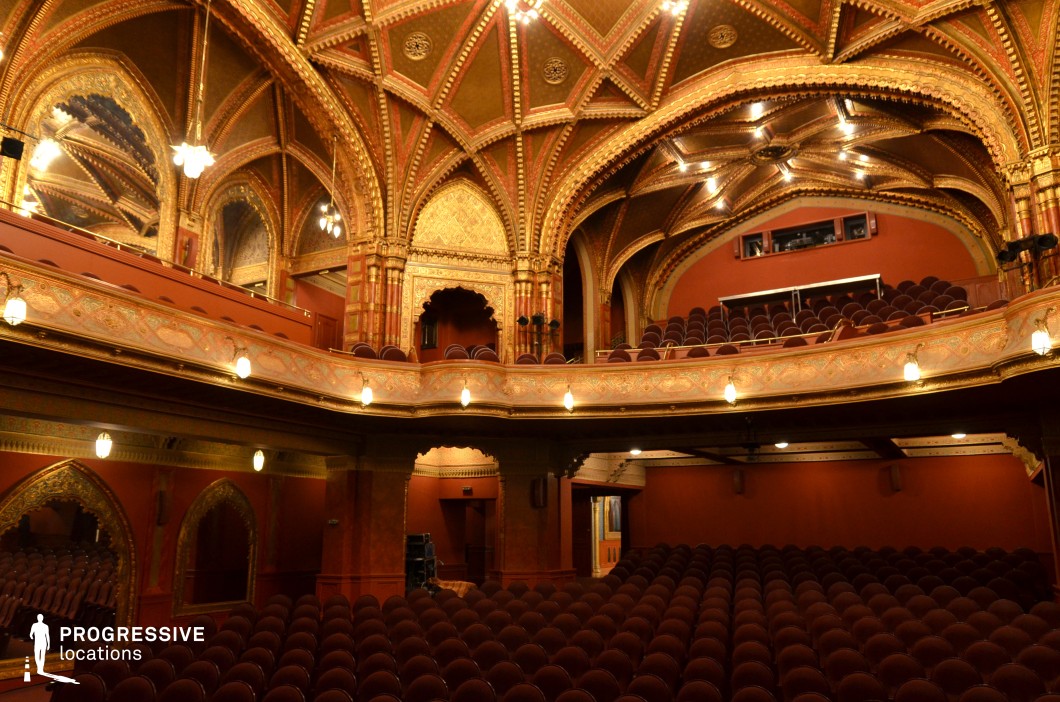 A breathtaking theater with intricate Moorish-inspired architecture, featuring pointed arches, gilded geometric patterns, and a richly decorated vaulted ceiling. Warm red and gold tones dominate the space, complemented by carved wooden details and ornate balcony railings. Rows of plush, dark red seats add to the luxurious ambiance, while soft, glowing lamps create an intimate, atmospheric lighting effect. The blend of historical grandeur and exotic ornamentation makes this venue ideal for period films, fantasy productions, or dramatic scenes requiring a regal and visually captivating setting.