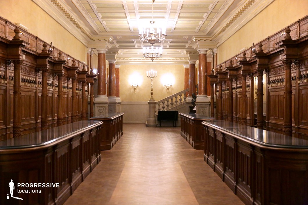 This lavishly designed cloakroom hall exudes old-world grandeur with its dark polished wood paneling, ornately carved columns, and reflective countertops. The symmetrical arrangement of wooden compartments and decorative moldings enhances the sense of order and elegance. The ceiling features intricate coffered detailing, while warm lighting from chandeliers and wall sconces creates a sophisticated ambiance. The space is framed by classical marble columns leading to a balustraded staircase, adding depth and visual interest. This location is ideal for period dramas, aristocratic settings, or historical thrillers requiring a regal and timeless atmosphere.