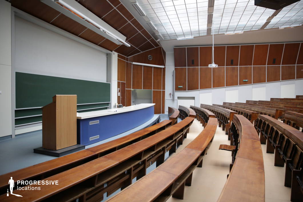 This traditional academic auditorium features curved wooden bench seating arranged in a semi-circular tiered layout, creating a classic lecture hall atmosphere. The wooden paneling on the walls and ceiling adds warmth, while the large glass-paneled ceiling allows natural light to illuminate the space. A sizable chalkboard and a curved podium with a blue front define the lecturer’s area, complemented by a suspended projector for modern presentations. The combination of vintage academic elements with functional updates makes this space ideal for university lectures, conferences, or formal educational settings.