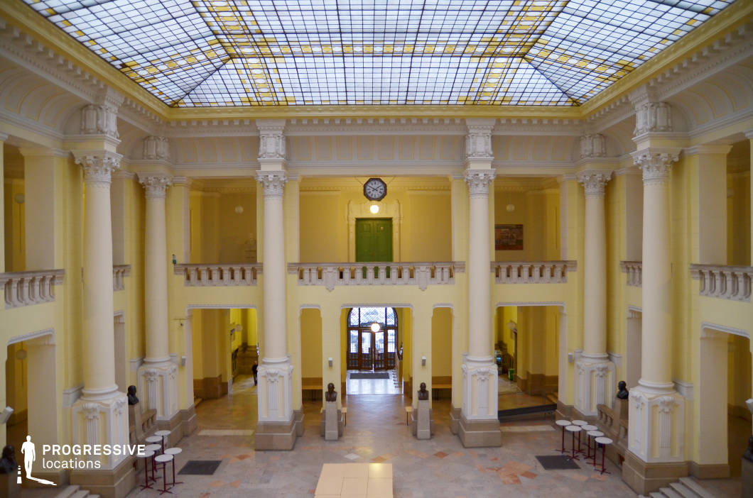 This grand neoclassical hall features towering Corinthian columns, intricate moldings, and a symmetrical layout that exudes elegance and prestige. The high glass-paneled ceiling, adorned with a geometric grid pattern, allows natural light to illuminate the space, enhancing the warm yellow-toned walls and polished stone flooring. A central balcony with balustrades overlooks the hall, adding depth and grandeur, while bust sculptures and period-style furnishings reinforce the academic and historical significance of the space. This setting is ideal for formal gatherings, institutional events, or cinematic scenes requiring classical sophistication.