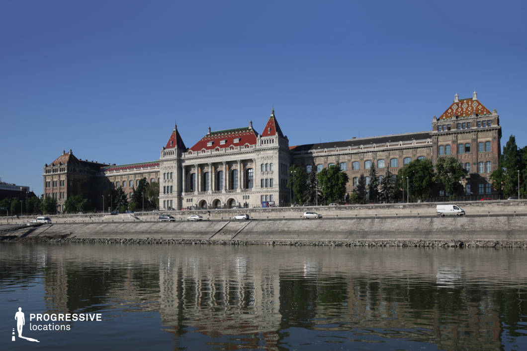 This imposing historic university building, located along a riverbank, showcases a blend of neogothic and neorenaissance architectural styles. The central structure is distinguished by its ornate facade, grand arched windows, and decorative stonework, while the red-tiled roofs with intricate patterns and corner turrets enhance its regal appearance. The symmetrical wings extend outward, featuring large rectangular windows and brickwork that evoke an academic and institutional ambiance. The riverside setting provides a scenic reflection of the building in the water, adding depth and visual interest. The prominent positioning along a waterfront embankment makes this an ideal location for scenes requiring a distinguished, intellectual, or European cityscape backdrop.