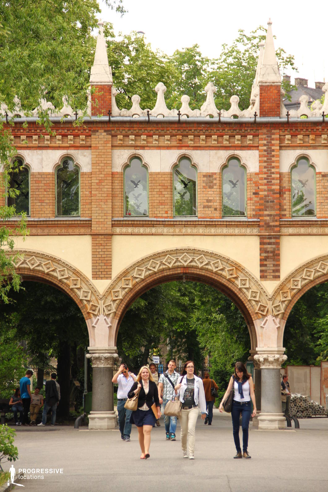 This striking neo-Gothic archway serves as a grand entrance to a university campus or historic institution. Constructed from warm-toned brick and accented with light stone detailing, the structure features pointed arches with intricate geometric carvings. Above the arches, a row of arched windows with decorative tracery adds to the historic charm, while ornamental pinnacles and crenelations create a castle-like silhouette. The archway frames a tree-lined pathway, enhancing the depth and inviting perspective of the scene. The combination of detailed craftsmanship and lush greenery makes this location ideal for settings requiring an academic, historical, or European architectural backdrop.
